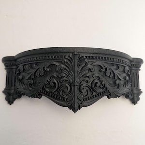 Könnte beinhalten: Ein schwarzes, verziertes Wandregal mit geschwungenem Design. Das Regal weist komplizierte, wirbelnde Muster und dekorative Säulen an jedem Ende auf. Das Regal besteht aus einem soliden Material und ist an einer weißen Wand befestigt.
