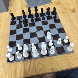 Peut inclure: Un jeu d'échecs noir et blanc avec les pièces disposées sur un échiquier à carreaux gris et noirs. Le plateau est composé de quatre carrés séparés qui peuvent être assemblés.