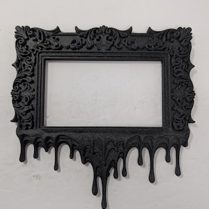 May include: Black rectangular picture frame with ornate detailing and a dripping effect at the bottom. The frame has a classic design with decorative swirls and curves, perfect for displaying photos or artwork.