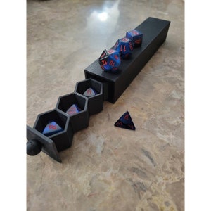 May include: A black hexagonal dice box with a sliding lid. The box is open and contains six blue dice with red numbers. One blue die with a red number four is outside the box.