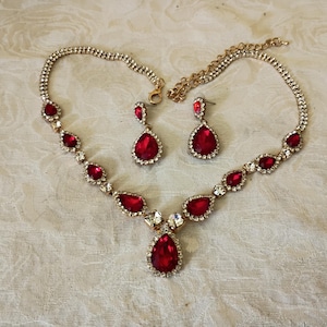 May include: A gold-tone necklace and earring set featuring teardrop-shaped red crystals surrounded by small clear rhinestones. The necklace has a central pendant and a chain with an adjustable clasp, while the earrings are matching teardrop drops.