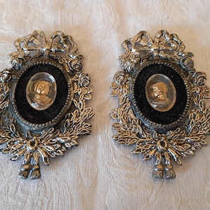 May include: Two ornate, silver-toned brooches. Each features an oval cameo set against black velvet within a decorative frame of leaves, bows, and floral accents. The cameos appear to be gold-toned, depicting a classical profile.