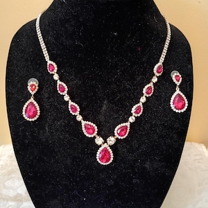 May include: A jewelry set consisting of a necklace and two matching earrings. The necklace features a series of teardrop-shaped red stones surrounded by small clear crystals, set in a gold-tone metal chain. Each earring has a single red teardrop stone.