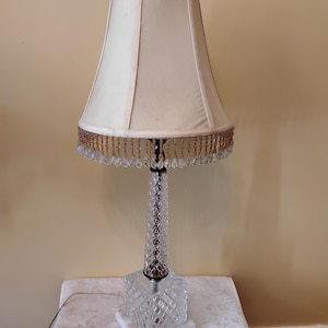 May include: A vintage table lamp with a cream-colored lampshade and a crystal base. The lampshade is trimmed with gold beads and crystal droplets. The lamp sits on a white marble base, adding a touch of elegance to the design.