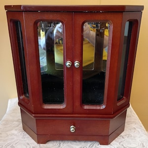 May include: A wooden jewelry armoire with a rich, reddish-brown finish. The octagonal cabinet features mirrored doors, a small drawer with a silver knob, and side panels. The armoire is set on a white patterned surface.