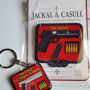 May include: Double-sided keychain featuring a black handgun design with the text "The Jackal" and "Jesus Christ Is In Heaven Now." The keychain also includes a design of bullets and a magazine. The second side features a silver handgun design with the text "Sanctified Anti-Vampiric Armament."