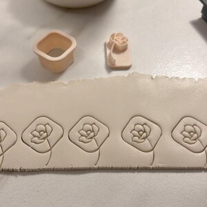 May include: A close-up of a white clay surface with four flower-shaped impressions made with a clay stamp. The stamp is visible in the background, along with a small square-shaped cutter.