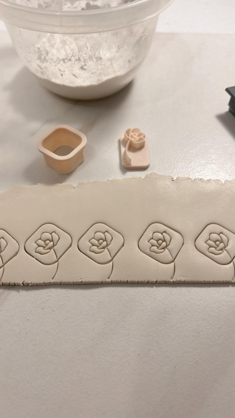 May include: A close-up of a white clay surface with four flower-shaped impressions made with a clay stamp. The stamp is visible in the background, along with a small square-shaped cutter.
