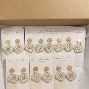 May include: Eight pairs of beige ceramic earrings with gold accents. Each earring features a floral design and is displayed on a white card with the text "Rosa Paulina Handmade Designs".