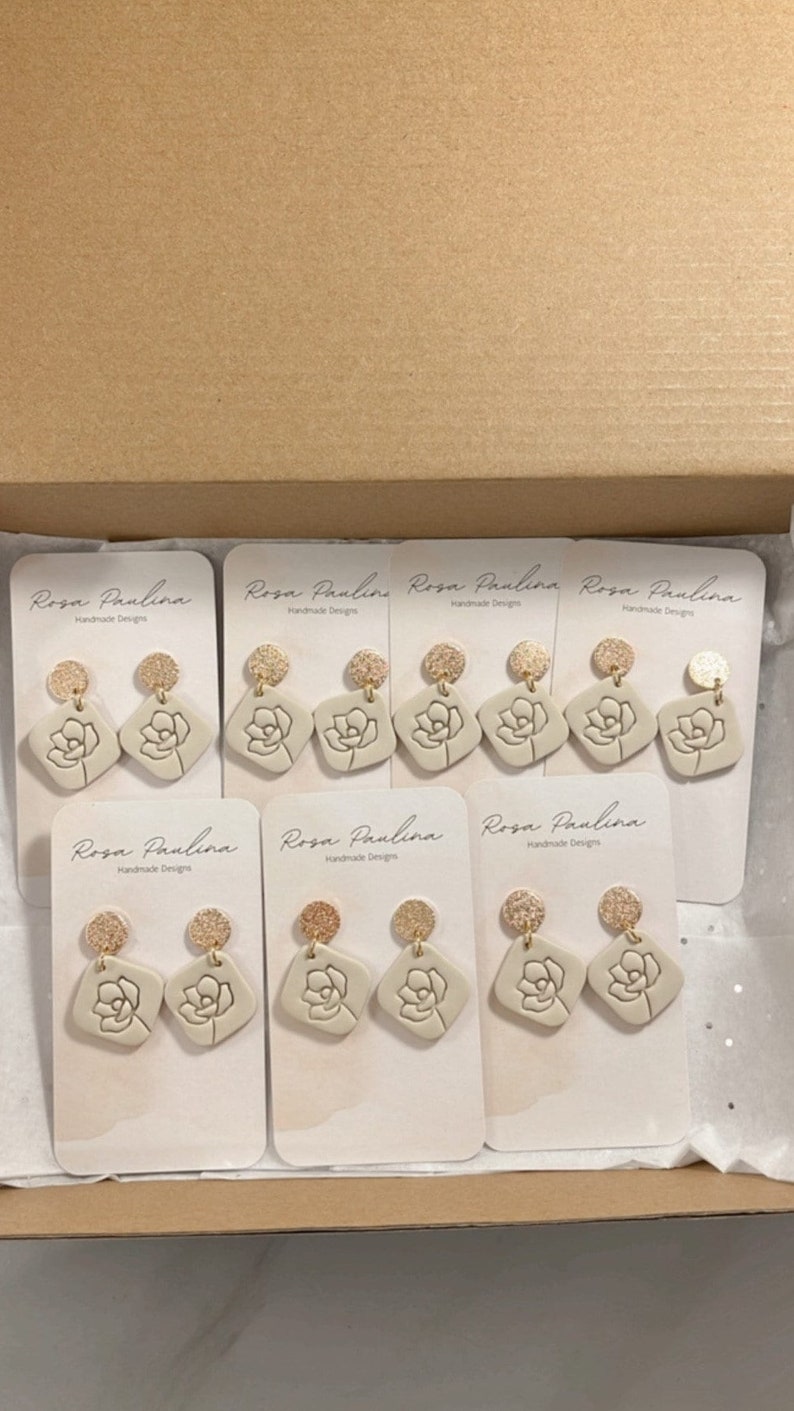 May include: Eight pairs of beige ceramic earrings with gold accents. Each earring features a floral design and is displayed on a white card with the text "Rosa Paulina Handmade Designs".