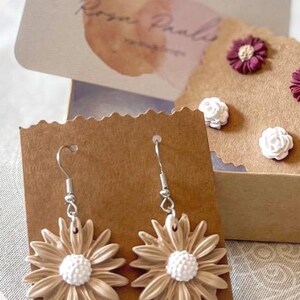 May include: A pair of dangle earrings featuring beige flower-shaped charms with white centers. The earrings are displayed on a brown card with the text "Rozen Pals" in white.