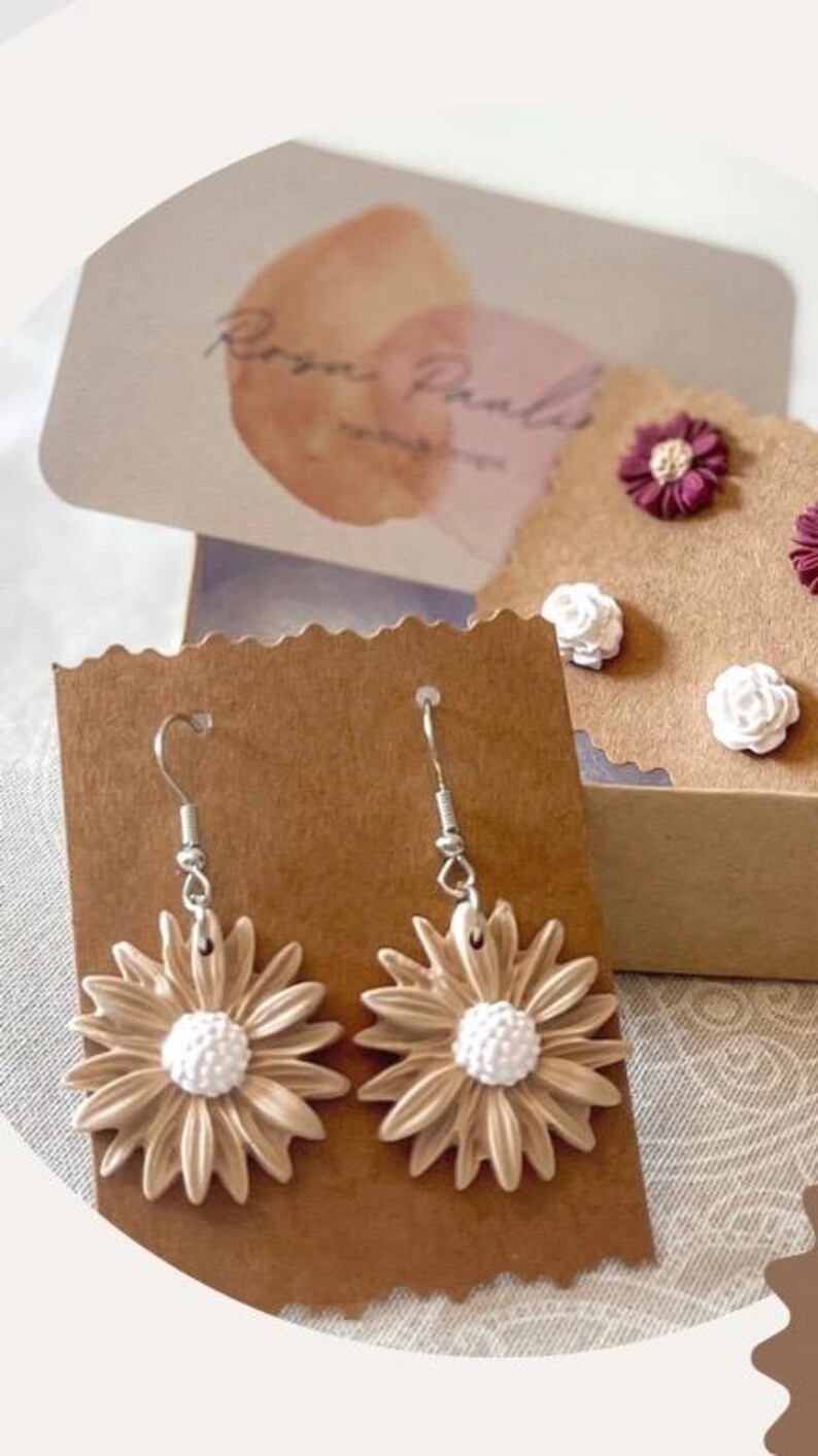 May include: A pair of dangle earrings featuring beige flower-shaped charms with white centers. The earrings are displayed on a brown card with the text "Rozen Pals" in white.