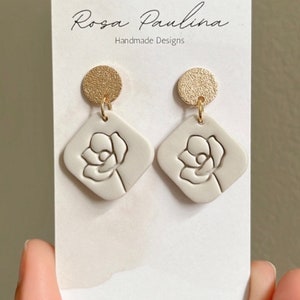May include: A pair of white ceramic earrings with a gold-toned disc and a raised flower design. The earrings are displayed on a white card with the text "Rosa Paulina Handmade Designs".