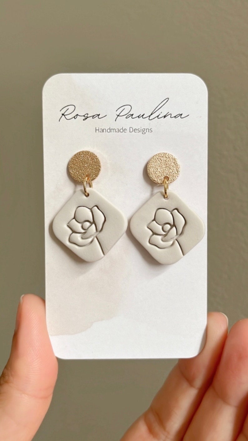 May include: A pair of white ceramic earrings with a gold-toned disc and a raised flower design. The earrings are displayed on a white card with the text "Rosa Paulina Handmade Designs".
