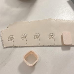 May include: A close-up of a white clay surface with four flower impressions. A white plastic square cutter and a white sponge are also visible.