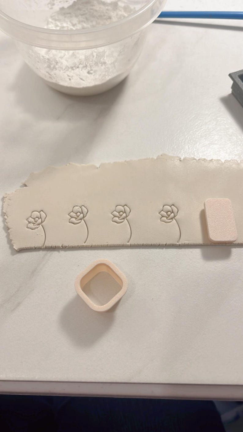 May include: A close-up of a white clay surface with four flower impressions. A white plastic square cutter and a white sponge are also visible.