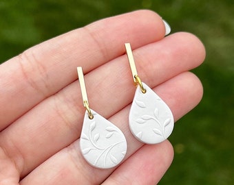 May include: A pair of white teardrop-shaped earrings with a delicate floral design. The earrings have gold posts and are made of polymer clay.