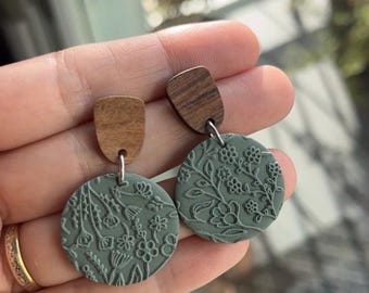 May include: A pair of handmade earrings featuring a wooden top and a circular, sage green clay pendant. The pendant is embossed with a floral pattern. The earrings are connected with a silver-colored metal loop.