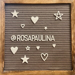 May include: A gray felt letter board with white letters spelling "@rosapaulina" surrounded by white stars and hearts.