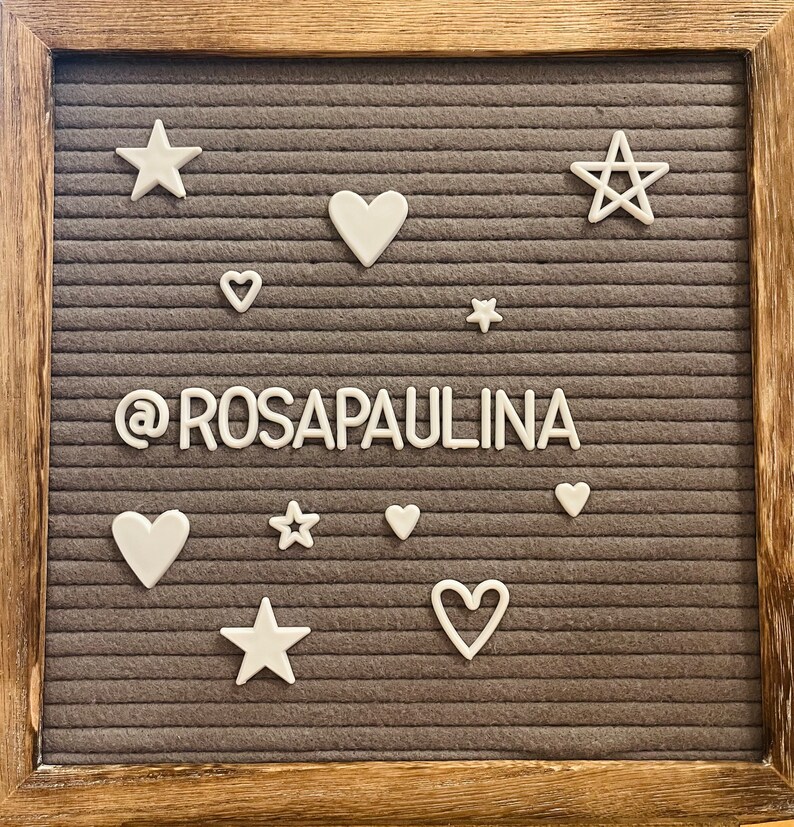 May include: A gray felt letter board with white letters spelling "@rosapaulina" surrounded by white stars and hearts.