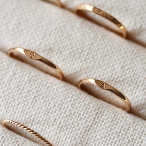 May include: A selection of gold rings, some with textured surfaces and others with engraved initials. The rings are presented on a light-coloured, textured background, highlighting their intricate designs and warm, metallic hues.