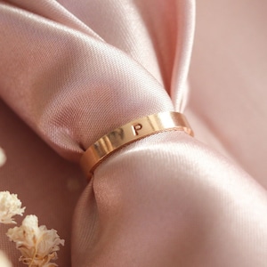 Puede incluir: Un anillo dorado con la letra "P" grabada en él, descansando sobre un fondo de tela rosa suave.