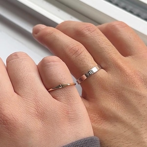 Puede incluir: Dos manos con anillos. Una mano tiene un anillo dorado con la letra "J", la otra un anillo plateado con la letra "R". Los anillos son bandas simples, con un fondo de ventana.