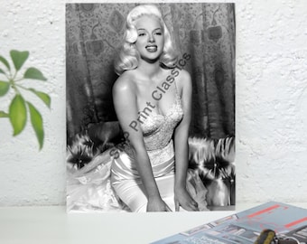 Könnte beinhalten: Schwarzweißfoto einer Frau mit blonden Haaren, die ein glitzerndes, tief ausgeschnittenes Kleid trägt. Sie sitzt auf einem Sofa mit einem gemusterten Stoff.