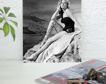 May include: Black and white photograph of a woman in a black swimsuit sitting on a rock. She is wearing a towel and has her hair styled in a glamorous updo. The background is a rocky landscape.