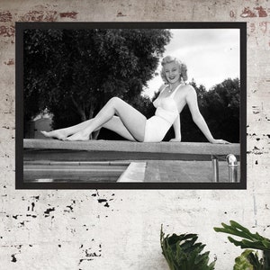 Puede incluir: Una fotografía en blanco y negro de una mujer en un traje de baño blanco sentada en un trampolín. Ella sonríe y mira a la cámara. La foto está enmarcada en un marco negro.