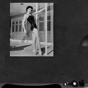 Könnte beinhalten: Schwarzweißfoto einer Frau im Badeanzug, die neben einem Pfosten steht. Sie lehnt sich an den Pfosten und schaut in die Kamera. Das Foto wurde im Freien aufgenommen und im Hintergrund ist ein Gebäude zu sehen.