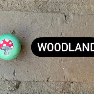 May include: A green painted rock with a red and white mushroom design. The text "WOODLAND" is written in white on a black oval.