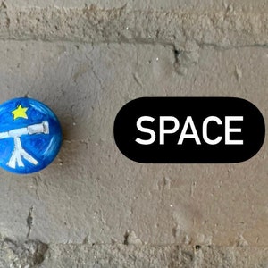 May include: A blue painted rock with a white telescope and a yellow star. The word "SPACE" is written in white on a black oval.