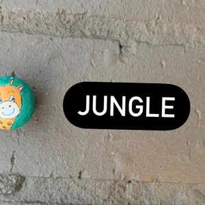May include: A painted rock with a green background and a cartoon giraffe face. The text "JUNGLE" is written on a black oval.