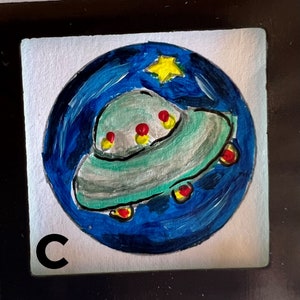May include: A hand-painted illustration of a flying saucer against a blue and black starry night sky. The saucer is green and gray with red and yellow accents. The image is framed in a black frame with the letter "C" in the lower left corner.