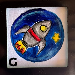 May include: A hand-painted tile with a blue and white circle depicting a rocket ship flying through space. The rocket ship is gray with red and yellow accents. A yellow star is visible in the background. The letter "G" is printed in black in the lower left corner of the tile.