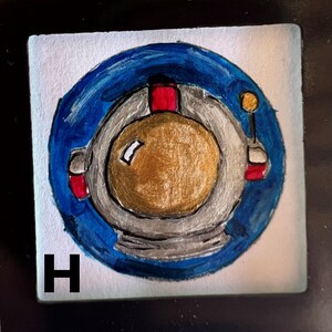 May include: A hand-painted tile with a blue circle containing a white astronaut helmet with a gold visor. The tile is labeled with the letter "H".