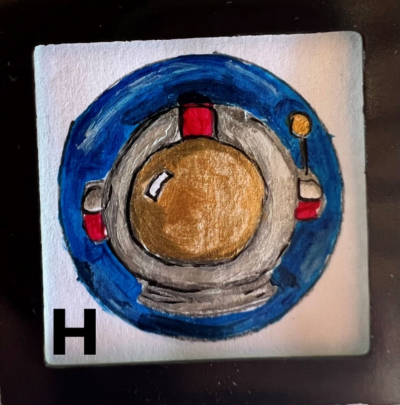 May include: A hand-painted tile with a blue circle containing a white astronaut helmet with a gold visor. The tile is labeled with the letter "H".