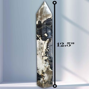 Puede incluir: Un obelisco de cristal alto y rectangular, de aproximadamente 31,75 cm de altura. La piedra pulida presenta un llamativo patrón de remolinos y vetas negras, blancas y marrones. La parte superior es puntiaguda y la forma general es simétrica.