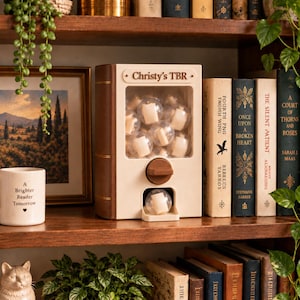 May include: A cream and brown book-shaped gumball machine dispenser with the text Christy's TBR printed on the top. The device contains multiple clear plastic spheres, each holding a small rolled paper scroll, with one sphere resting in the dispenser tray.
