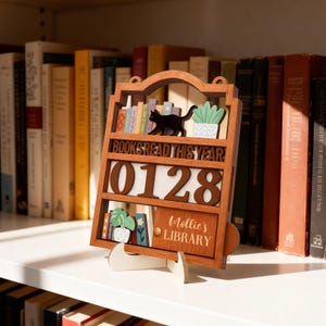 May include: A wooden bookshelf-style calendar displaying "BOOKS READ THIS YEAR" and the numbers "0128". It includes a black cat, miniature books, and potted plants. The bottom shelf reads "Mollie's LIBRARY". The calendar is on a white stand.