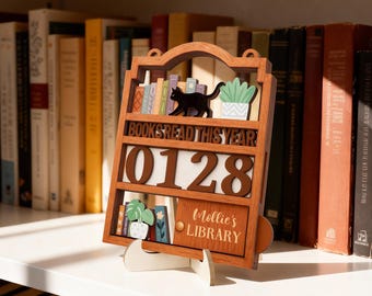 May include: A wooden bookshelf-shaped calendar with the text "BOOKS READ THIS YEAR" and the numbers "0128". It features a black cat figurine, miniature books, and potted plants. The bottom shelf reads "Mollie's LIBRARY".