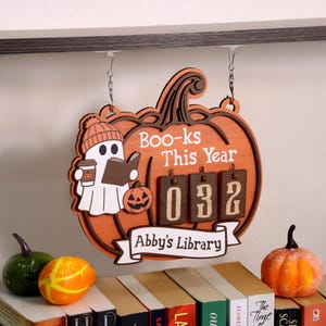 Peut inclure: Panneau en bois en forme de citrouille avec le texte "Boo-ks This Year" et "Abby's Library". Un fantôme avec une tasse de café et un livre est à gauche. Le panneau a un compteur affichant "032". Des citrouilles décoratives et des livres sont au premier plan.