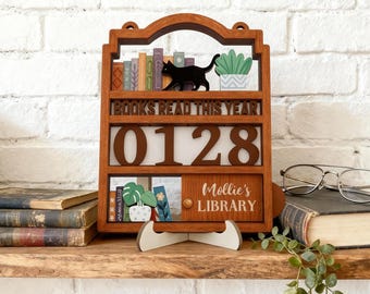 May include: A wooden bookshelf-style display with the text "BOOKS READ THIS YEAR" and the number "0128". It features a black cat silhouette, miniature books, and potted plants. The bottom section reads "Mollie's LIBRARY".
