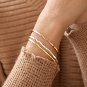 Puede incluir: Tres brazaletes finos de metal grabado en oro rosa, plata y oro están apilados en una muñeca. Los brazaletes tienen frases inspiradoras grabadas. La persona lleva un suéter marrón.