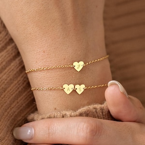 Peut inclure: Deux bracelets dorés délicats avec des breloques en forme de cœur. L'un des bracelets comporte un seul cœur avec la lettre "J", tandis que l'autre en a deux avec les lettres "A" et "S". Les bracelets sont portés sur un poignet.