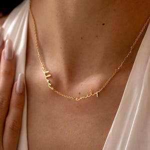 May include: A fine gold necklace with the names "Melissa" and "emily" in a cursive script. The necklace is shown on a person wearing a white top, emphasising the design and colour of the jewellery.