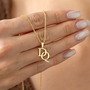 Peut inclure: Un collier doré avec un pendentif représentant les lettres entrelacées "DQ". Le pendentif et la chaîne sont délicats, et le collier est présenté sur un fond flou. Le bijou est probablement en métal plaqué or.