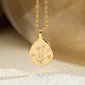 Op de afbeelding: Een goudkleurige hangerketting met een druppelvormige bedel. De bedel heeft een gegraveerd bloemenontwerp. De ketting en hanger zijn gemaakt van glanzend, goudkleurig metaal. De achtergrond is een zachte, neutrale kleur.