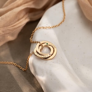 Puede incluir: Un collar de tono dorado con tres colgantes circulares entrelazados. Los anillos están grabados con el texto MMXXIV y 52.52° N, 13.405° E. Un anillo tiene una pequeña piedra verde y otro cuenta con una pequeña piedra de color violeta.